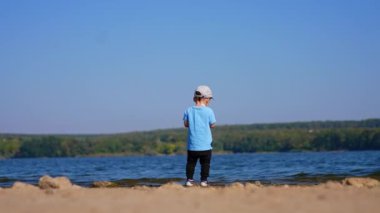 Rıhtımda duran bir bebeğin arka görüntüsü. Küçük çocuk taşları suya atıp yenilerini seçiyor..