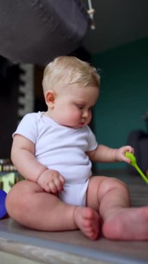 Beyaz elbiseli sevimli bebek yerde oturuyor. Sevimli bebek mutfak silikon fırçasıyla oynuyor. Dikey video.
