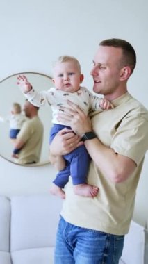 Babası tatlı bebeğini aynanın karşısında tutuyor. Bebek elini sallarken yana bakıyor. Dikey video.