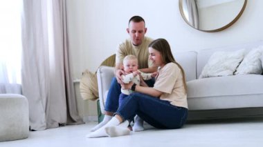 Long-haired Caucasian woman sits on the floor near the sofa supporting her son. Man sits behind on the sofa looking at his wife and baby. Low angle view.