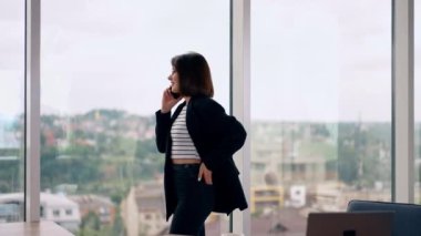 Genç bir kadın panoramik pencerenin önünde telefon konuşması yapıyor. Ofiste iş görüşmesi var..