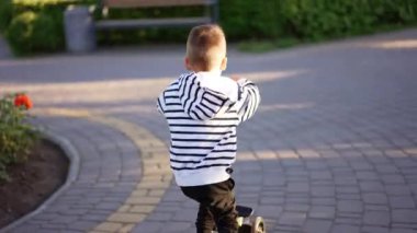 Parkta scooterını süren küçük bir çocuğu takip etmek. Beyaz ve siyah çizgili kazaklı çocuk dışarıda eğleniyor..