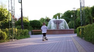 Çizgili tişörtlü çocuk fıskiyeye doğru scooterını sürüyor. Parkta eğlenen bir çocuğun arka görüntüsü..