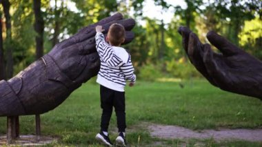 Parktaki büyük ahşap ellerin heykelinin önünde duran üç yaşında bir çocuk. Çocuk hem heykelleri hem de yaprakları kontrol ediyor..