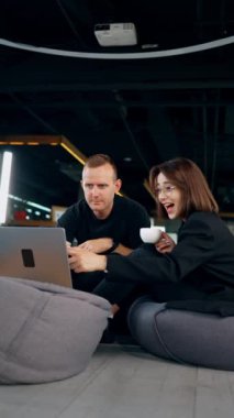 Birlikte çalışan ve neşeli iletişim kuran insanlar. Erkek ve kadın laptopa odaklanmış bir şekilde fasulye çuvalında oturuyorlar. Dikey video.