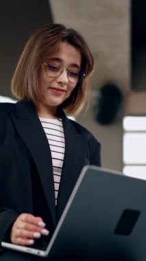 Gözlük takan güzel bir esmer dizüstü bilgisayarda çalışıyor. Uzaktan kumandayla çalışan kendine güvenen modern bir kadının portresi. Düşük açı. Dikey video.