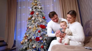 Annenin dizlerinin üzerinde oturan küçük bebek oyuncağına odaklanmış ve onu ısırmaya çalışıyor. Ebeveynler çocuklarına sevgiyle bakar. Arkaplanda süslenmiş Noel ağacı.