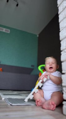 Güzel, tombul, sarı saçlı erkek bebek yerde oturuyor. Küçük çocuk diş çıkarıyor. Ağzına bir şeyler çek. Dikey video.