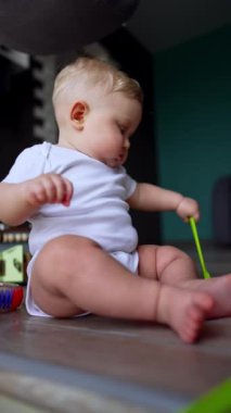 Beyaz tulumlu, açık renk saçlı bebek yerde oturuyor. Küçük çocuk mutfak fırçasıyla oynuyor. Dikey video.