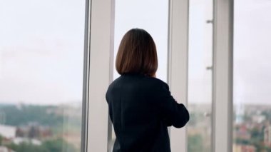 Pencerede duran esmer bir kadının arka görüntüsü. Gülümseyen kadın telefonda konuşurken kameraya dönüyor..