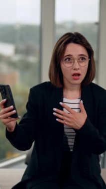 Çekici, koyu renk saçlı bayan telefonuna hayretle bakıyor. Kadın aletini ağzından düşmüş bir şekilde gösteriyor. Dikey video.