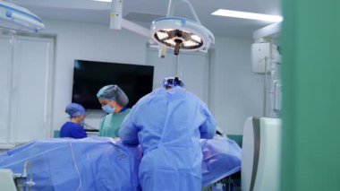 Ameliyat masasına yaslanmış bir cerrahın arka görüntüsü. Hemşire hastanın diğer tarafında duruyor ve kullanılmış aletleri temizliyor. Doktor yan tarafa bakıyor ve kameraya doğru dönüyor..