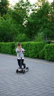Neşeli Kafkas çocuk scooterının yanındaki parka doğru koşuyor. Yeni yürümeye başlayan mutlu çocuk dışarıda eğleniyor. Dikey video.