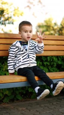 Dışarıdaki bankta oturan şirin beyaz bir bebek. Çocuk yaşını parmaklarıyla gösteriyor ve ayaklarını sallıyor. Dikey video.