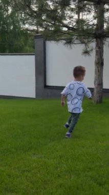 Yeşil çimlerin ve asfalt yolların orada koşan neşeli beyaz bir oğlan. Aktif küçük çocuk dışarıda eğleniyor. Dikey video.