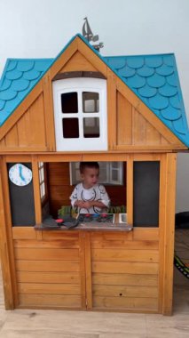 Kafkasyalı küçük çocuk ahşap oyun evinde oynuyor. Çocuk evden çıkıyor ve köşeden dikizliyor. Üst Manzara. Dikey video.