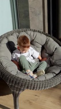 Küçük çocuk dışarıdaki yuvarlak koltukta oturuyor. Küçük çocuk ayağa kalkar ve kapıya yönelir. Dikey video.