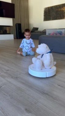 Tatlı çocuk, dönen robot elektrikli süpürgenin üzerinde oturan büyük beyaz ayıcıkla ilgileniyor. Çocuk gülerek cihazın çalışmasını izliyor. Dikey video.