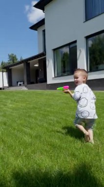 Avluda elinde su tabancasıyla çimlerin yanından koşan enerjik beyaz çocuk. Mutlu çocuk yazın dışarıda eğleniyor. Dikey video.