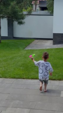 Küçük çocuk elinde su tabancası olan yeşil çimlerle avluya koşuyor. Mutlu çocuk yazın evinin yakınındaki çimenlikte oynuyor. Dikey video.