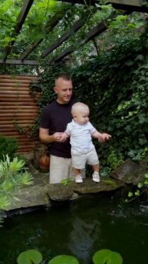 Baba küçük bebeğini fıskiyenin yanında tutuyor. Suyun akışına bakan tatlı bir çocuk. Arkaplanda güzel bir yeşillik. Dikey video.