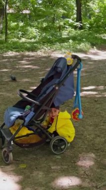 Parlak sarı ceket giyen çocuk bebek arabasının yanında oturup bir şey arıyor. Çocuk ayağa kalkar, kaçar ve önüne bir şey atar. Dikey video.