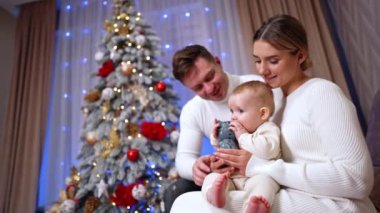 Güzel, beyaz bir bebek annesinin elinde bir oyuncak tutuyor ve onu ısırmaya çalışıyor. Babam arkada oturup oğluna bakıyor. Ailesinin Noel 'de birlikte zaman geçirmesine düşük açılı bakış açısı. Dikey video.