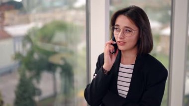 Güzel pozitif bayan pencerede durup telefonda konuşuyor. Ofisteki müşterilerle ya da müşterilerle konuşmak. Bulanık arkaplan.