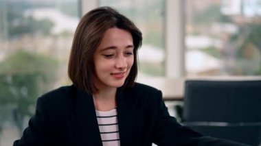 Attractive smiling Caucasian brunette lady sits indoors. Woman puts on her glasses and starts work. Close up. Blurred backdrop.