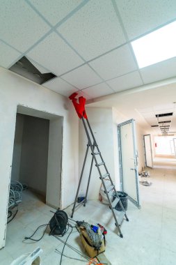 Ofis koridorunun tadilatında tavan kiremitlerini tamir eden işçi. Kırmızı tulumlu bir işçi kısmen yenilenmiş bir ofis koridorundaki merdivenin tavan fayanslarını tamir ediyor..