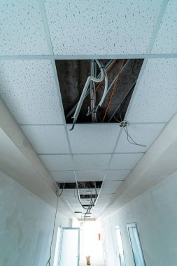 Açık kablolar ve aydınlatma armatürleriyle dolu bir koridorda tavan tamiri. İşçiler bir koridor tavanını onarıyor, yukarıdaki elektrik tesisatlarını ve elektrik tesisatlarını açıyorlar.