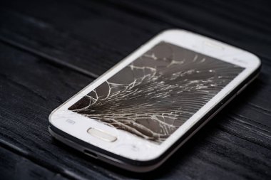 Koyu ahşap bir yüzeyde çatlamış bir akıllı telefon. Bir akıllı telefon koyu ahşap bir yüzeye dayanır, ekranı görünür çatlaklarla parçalanır.