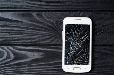 Ahşap bir masada karanlık bir kapanışla duran kırık bir akıllı telefon. Zarar görmüş beyaz bir akıllı telefon parlak siyah bir ahşap masanın üzerinde duruyor.
