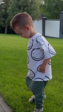 Sevimli beyaz çocuk çimlerin üzerinde yürüyor ve etrafında dönüyor. Bebek yazın dışarıda çok güzel vakit geçiriyor. Dikey video.