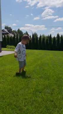 Güneşli bir yaz gününde çimlerin üzerinde koşan çıplak ayaklı beyaz çocuk. Aktif erkek bebek dışarıda enerjik bir şekilde koşuyor. Dikey video.