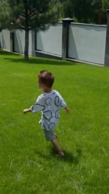 Neşeli beyaz çocuk yeşil çimlerin üzerinde hızla koşuyor. Aktif erkek bebek yazın dışarıda kalıyor. Dikey video.