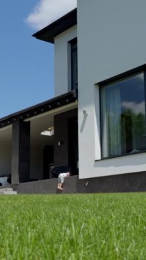 Küçük oğlan büyük bir evin verandasına tırmanıyor. Çocuk merdivenlere gidiyor ve kameraya yaklaşan yeşil çimlerin yanından geçiyor. Dikey video.