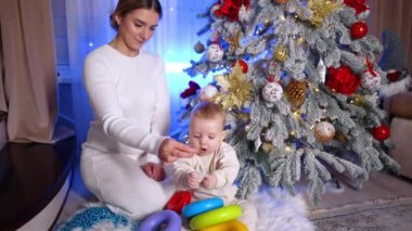 Beyaz elbiseli çekici bir kadın Noel ağacında bebeğiyle oturuyor. Anne oğluna kar tanesi oyuncağı süslemesi gösteriyor..