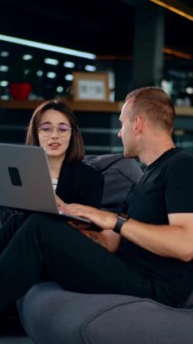 Modern ofiste çalışan meslektaşlar sohbet ediyorlar. Kadın ve erkek fasulye koltuklarında oturup laptopa bakar ve konuşurlar. Dikey video.