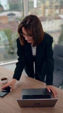 Kadın dizüstü bilgisayarına yaslanmış duruyor. Siyah ceketli kadın telefonunu alır ve pencereye gider. Dikey video.