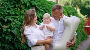Sevgi dolu beyaz anne babalar bebeklerini kafasından öperken tutarlar. Adam elinde bir numara şeklinde bir balon tutuyor. Arkaplanda yeşillik.