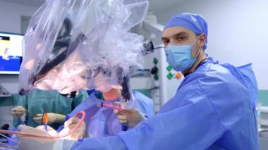 Bir şırınga tutan asistan mikroskoba arkasını döner ve kameraya bakar. Nöroşirürji ameliyatının ilerlemesi.