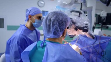 Hastayı ameliyat eden ve mikroskoba bakan bir doktorun arka görüntüsü. Erkek asistan hastanın yanına oturur..