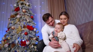 Kanepede oturan üç kişilik güzel bir beyaz aile. Anne ve babalar güzel oğullarını bir oyuncakla oynarken kucaklar. Arkaplanda Noel ağacı.