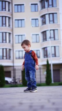 Beyaz, tatlı çocuk dışarıda sabun köpüğüyle oynuyor. Kot pantolonlu çocuk onları yakalamak için köpüklerle koşuyor. Düşük açı. Dikey video.