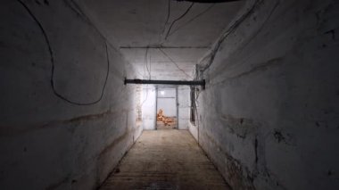 Çıplak duvarları ve tavandan sarkan kabloları olan uzun bir koridorda hareket. Ortasında tuğla duvar olan iyi ışıklandırılmış odaya girmek..