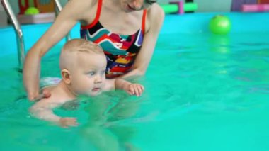 Güzel sarışın erkek bebek bez takıyor ve yüzme öğreniyor. Tanınmayan koç havuzda bir çocuğu tutuyor..
