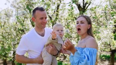 Beyaz ebeveynler ve küçük oğulları bahçede dikiliyorlar. Anne oğluna bir karahindiba gösteriyor. Aileler çiçeğe üflüyor..