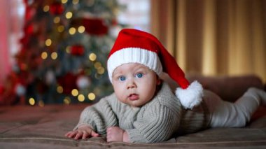 Büyük mavi gözlü sevimli beyaz bebek kameraya bakarak yatıyor. Güzel bebek Noel Baba şapkası ve kalın kazak giyiyor. Arkaplanda bulanık Noel ağacı.