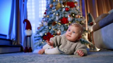 Sevimli küçük bebek sıcak kazak giyip yerde yatıyor. Tatlı sarışın çocuk halıda sürünüyor. Bulanık arkaplanda Noel ağacı.